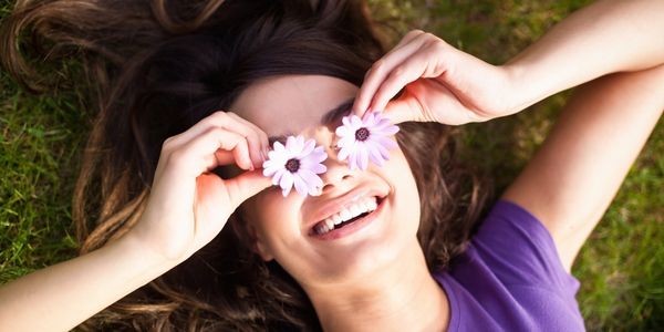 Smiling young woman laying down on the grass with flowers on her eyes instead of sunglasses.