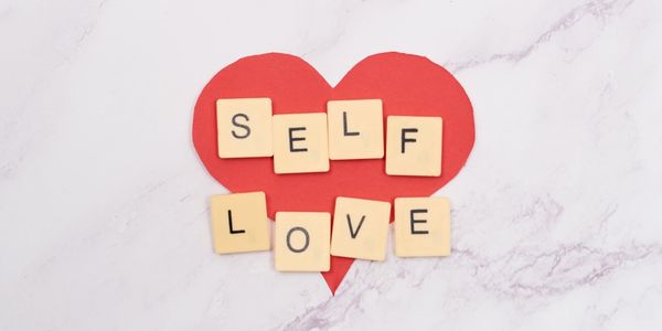 Self-love is spelled out with lettered squares on top of a red love heart cardboard cut out.