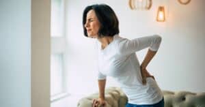 Adult woman is holding her lower back, while standing and suffering from unbearable chronic pain
