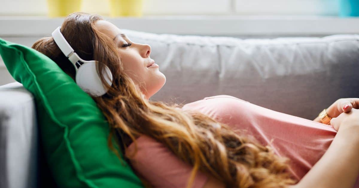 young woman lying on couch with her eyes closed, wearing headphones practicing binaural beats meditation and looking calm and peaceful